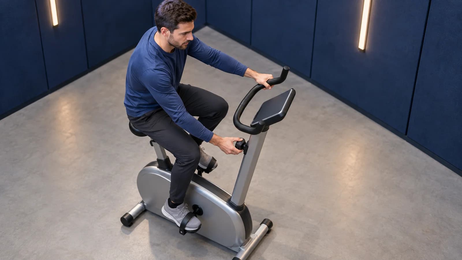 Männlicher Anfänger tritt auf einem Ergometer und dreht in einer indigoblauen Cardio-Nische am Widerstandsregler.